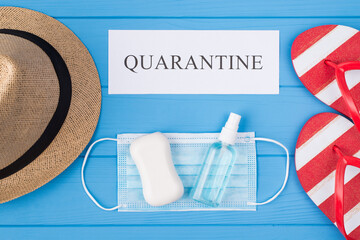 Quarantine and summertime fun concept. Top above overhead view photo of soap sanitizer mask flipflops hat and word quarantine isolated on blue wooden background