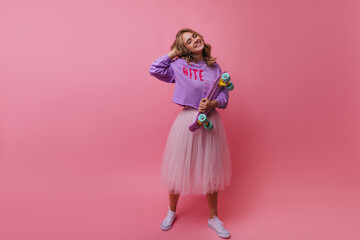 Full-length shot of graceful girl in long lush skirt holding skateboard. Indoor photo of smiling romantic blonde woman with longboard. © Look!