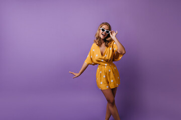 Winsome tanned girl in sunglasses posing with excited smile. Blonde carefree woman in yellow attire dancing on purple background.