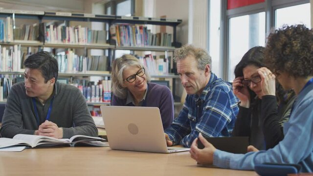 Female teacher helping group of mature multi-cultural students studying and working on laptop in library learning IT skills in evening class - shot in slow motion