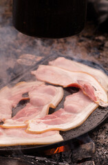 Middle bacon rashers cook on an iron plate, over an open campfire - smoke and steam fill the chilly morning air - with a black billy hanging above. 