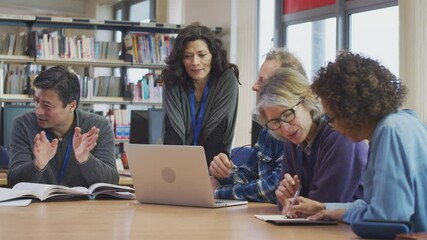 Female teacher helping group of mature multi-cultural students studying and working on laptop in library learning IT skills in evening class - shot in slow motion - Powered by Adobe