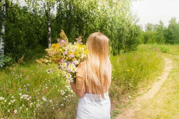 A beautiful European girl with a large bouquet of wild flowers stands by the road in the forest on a Sunny summer day.