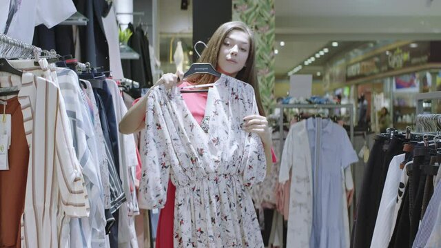 Young pretty woman is trying floral printed dress while standing between clothes racks in the shop. Attractive girl is looking at herself to the camera like in mirror, turning and smiling.