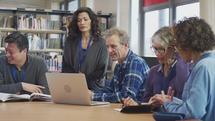 Female teacher helping group of mature multi-cultural students studying and working on laptop in library learning IT skills in evening class - shot in slow motion - Powered by Adobe