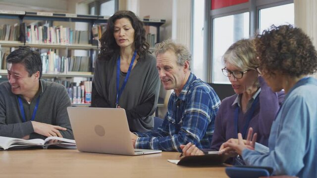 Female teacher helping group of mature multi-cultural students studying and working on laptop in library learning IT skills in evening class - shot in slow motion