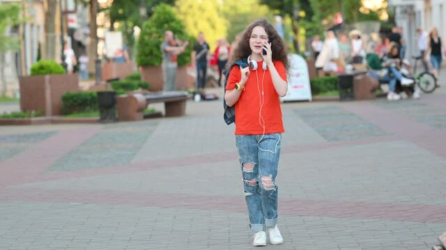 Young Attractive Girl Walking Street City Communicates on Smartphone. Girl with Headphones on Her Neck Uses Mobile Talking and Smiling.