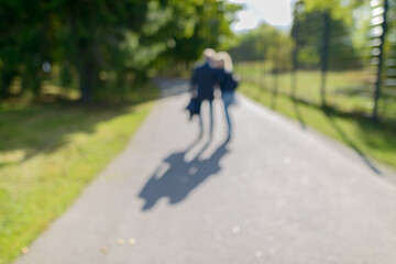 Obraz premium Defocused couple walking in the park along path of green lush trees on sunny day