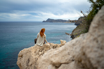 Rear view of woman sitting on rock by sea against cloudy sky