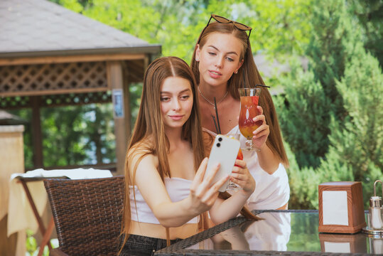beautiful, young girls with fruit cocktails take a selfie on their smartphone in a summer cafe against the background of nature