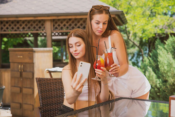 beautiful, young girls with fruit cocktails take a selfie on their smartphone in a summer cafe against the background of nature
