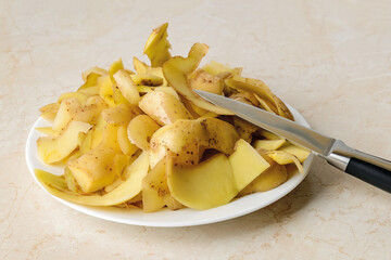 Heap of fresh potato peels and small vegetable knife on a white plate ober kitchen table. Cook boiled potatoes at home. Household biodegradable waste.