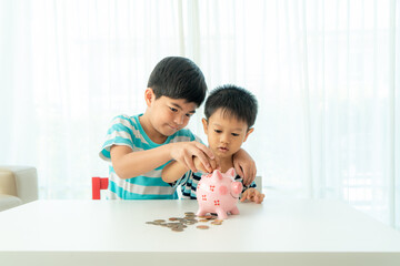 Two Asian cute brother and son insert coin  in piggy bank together on the table in living room at home. Concept of bonding of sibling, friendship and saving, investment money concept