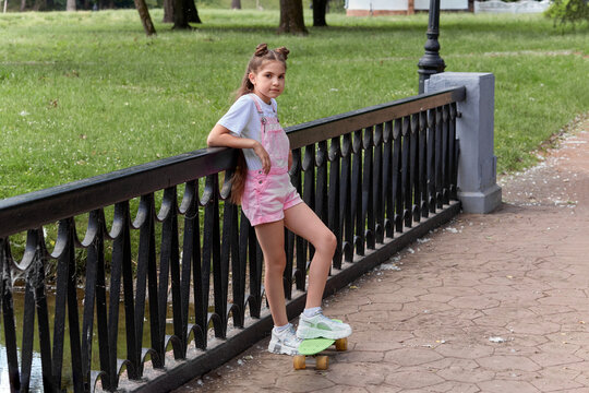 Young beautiful cute girl with long hair stands on a skateboard near the fence.