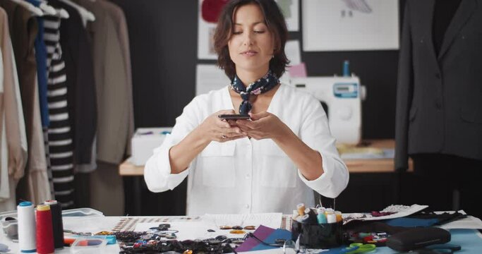 Attractive female designer in white shirt taking photos of sketches on modern smartphone. Dark-haired woman using digital gadget at workshop studio.