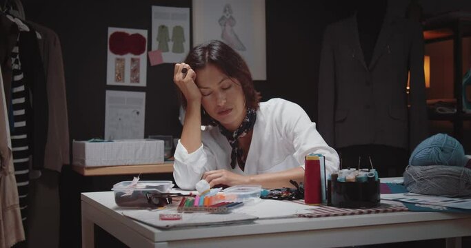 Tired fashion designer with brown hair fell asleep at tailor studio while working overtime. Mature woman sitting at table with dark atmosphere around.