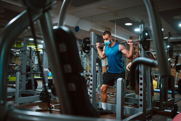 A young caucasian athlete man with a mask on his face exercises and lifts weights in the gym. COVID...