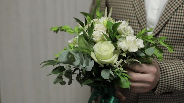 Groom with wedding bouquet in his hands at home. White shirt, jacket. Close-up shot. Slow motion
