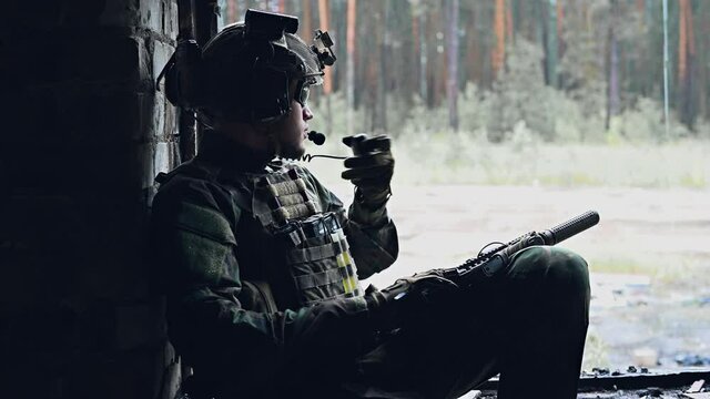 Military staging base, soldier uses walkie-talkie radio while sitting in destroyed building and resting after operation. Raining outside in a forest.