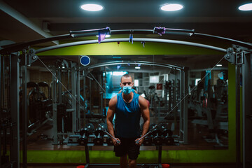 Portrait od young muscular fitness man with medical mask doing heavy weight exercise for body on machine in the gym.Prevention of COVID - 19 coronavirus