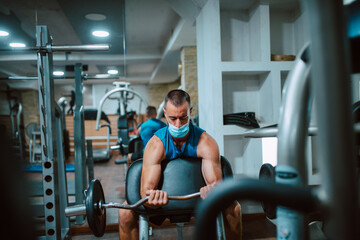 A young caucasian athlete man with a mask on his face exercises and lifts weights in the gym. COVID 19 coronavirus protection