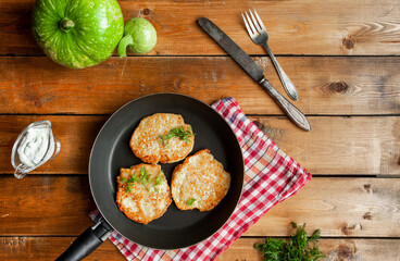 zucchini fritters on a black small pan, red checkered napkin, silver vintage Cutlery, green zucchini, greens, sauce bowl with white sauce on a wooden table, top view, simple village food