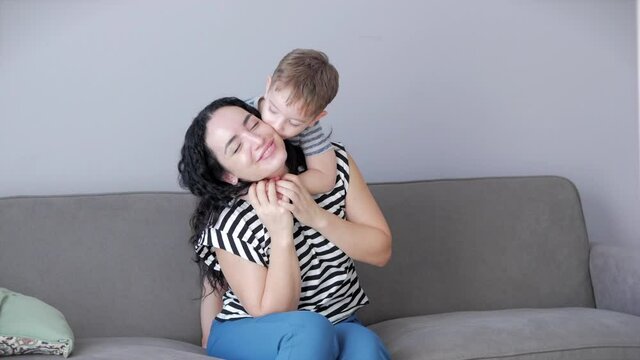 Happy healthy family young mom hugs cute little child son who kisses mother on lips or cheek sitting on sofa at home, funny son kid boy love games mother.