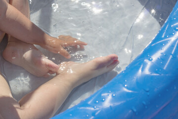 Feet and hand of a child bathing in a blue plastic pool. Little kid plays in a transparent pool with dandelions. Water procedures in the air. Beautiful shadows from a dandelion, the sun in the water