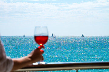 The human hand with rose wineglass on a balcony railing on backgroun of the seascape with sail yachts on a horizon, focus on background.