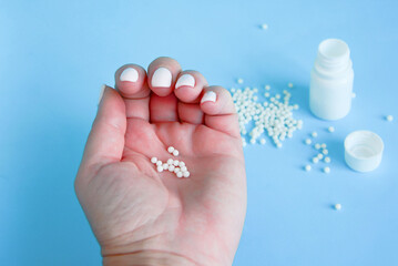 Woman's hand with homeopathic pills  globules and white plastic bottle on blue background. Alternative Homeopathy medicine herbs, healtcare and pills concept. 