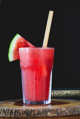 A fresh watermelon smoothie served with a bamboo straw on wood with glass bottle and black background 