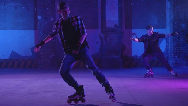 Duet of two young men synchronously skating professionally on stage under bright pink and blue lighting and braking at the same time, side view with focus on the guys. Cool performance from skaters.