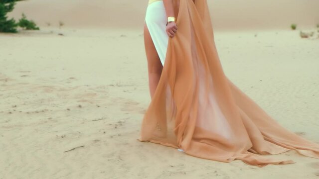 Closeup portrait beautiful female legs walking, hot summer beach, Egyptian desert. Beauty bare feet. Gold jewelry for legs profiles, payals, bracelets. White long dress, orange cloak. Trees, pyramids