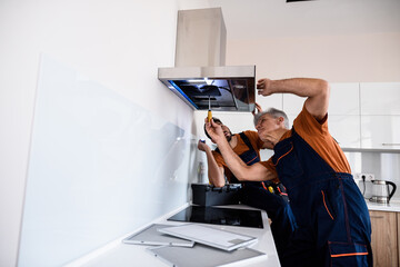 Two workers, handyman in uniform installing or repairing a kitchen extractor, replacing filter in...
