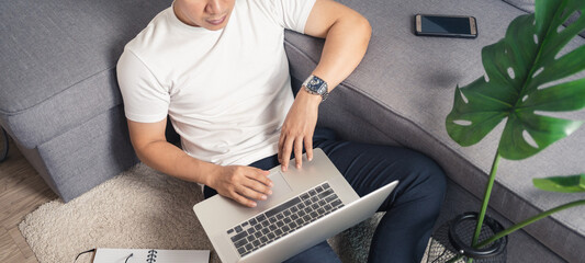 Close up of young Asian man sitting on floor working from home using laptop computer and notebook. Work from home concept. Banner portion