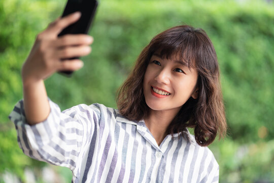 Young beautiful Asian woman using smartphone taking selfie photo. A happy girl enjoying outdoor lifestyle surrounded by greeny plant. Positive thinking concept