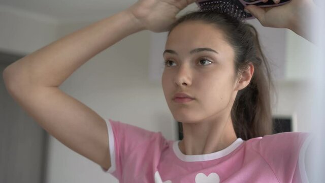 tender schoolgirl brushes long hair in ponytail with black hairbrush standing at large mirror close view