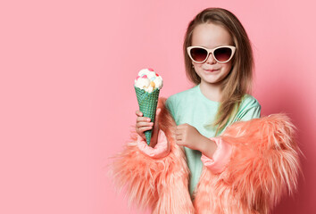 Glad and smiling kid girl in faux fur coat and sunglasses holds big ice cream with candies in...