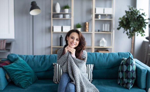 Young woman snuggling into a warm blanket sitting on a cozy sofa at home. Female portrait.