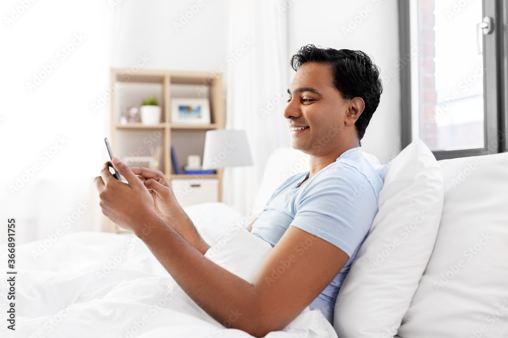 Wall mural technology, bedtime and rest concept - happy smiling indian man with smartphone lying in bed at home