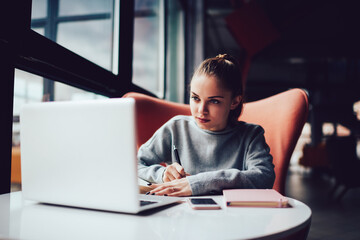 Beautiful female student preparing for university examination learning information online and making notes.Skilled marketing expert pondering on creative solution and making plans analyzing content