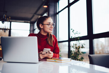 Positive pensive female blogger looking away while chatting with followers in personal website via smartphone.Dreaming cute hipster girl in eyewear sending messages to friends using modern device