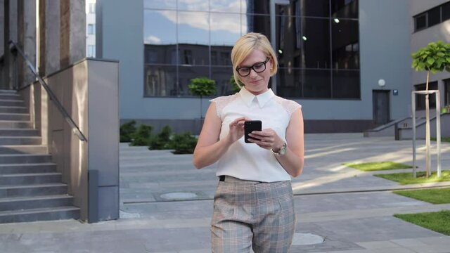 Blonde businesswoman looks away and then looks at a mobile phone and flips through the news using a smartphone. Stylish clothes, glasses, luxurious look.