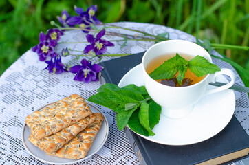 A Cup of green tea with a mint leaf, flowers, and a book on a table in the garden. Evening tea with cookies. Summer village concept. High quality photo