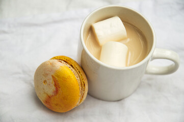 Close up, macro. A cup of hot latte with marshmallows, orange macaron next to it.