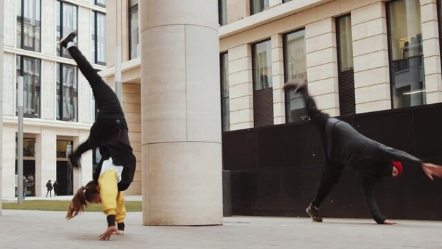 Slow motion shot of male and female parkour athletes performing cartwheel and backflips in a row while practicing tricks together outdoors in the city