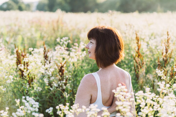 Fototapeta na wymiar Middle aged mature red haired woman in white top with sensitive skin and freckles during yoga routine in sunny morning enjoys sunset in summer meadow field with chamomiles flowers.