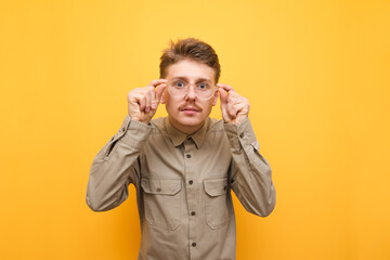 Fototapeta premium Portrait of a funny nerd with glasses and mustache on a yellow background, looks into the camera with a serious face and adjusts his glasses.Serious geek in his shirt looks intently into the camera