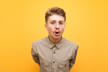 Portrait of surprised young nerd man on yellow background, looks surprised at camera, wears beige shirt and glasses for vision. Isolated. Copy space