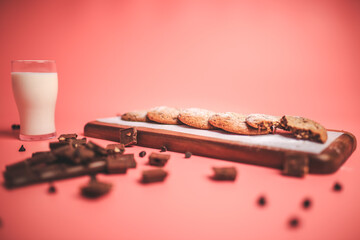 Freshly made Chocolate Chip Cookies on a wooden serving board. Cookies with a glass of fresh milk on the side along with chocolate chips and pieces of chocolate around on a pink background.Freshly mad